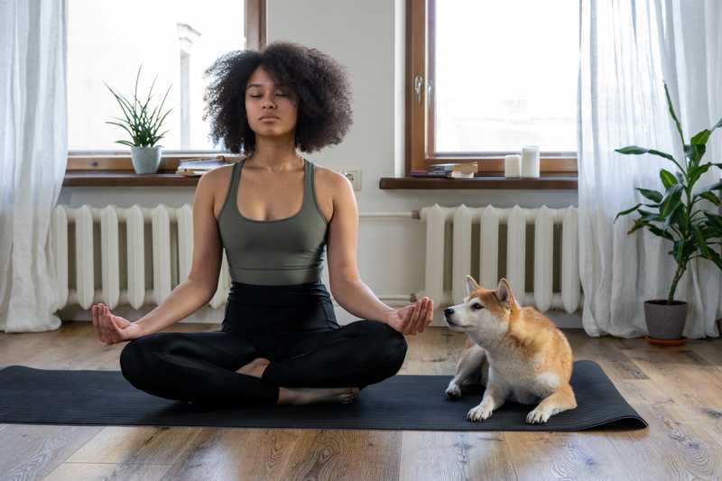 Woman and dog sit on yoga mat together while woman meditates